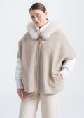 Woman wearing a beige alpaca cape with light beige real fur trimmed hood over a separate white quilted puffer jacket, showing ribbed cuffs, gold front zipper and discreet side welt pockets by Lebeni.