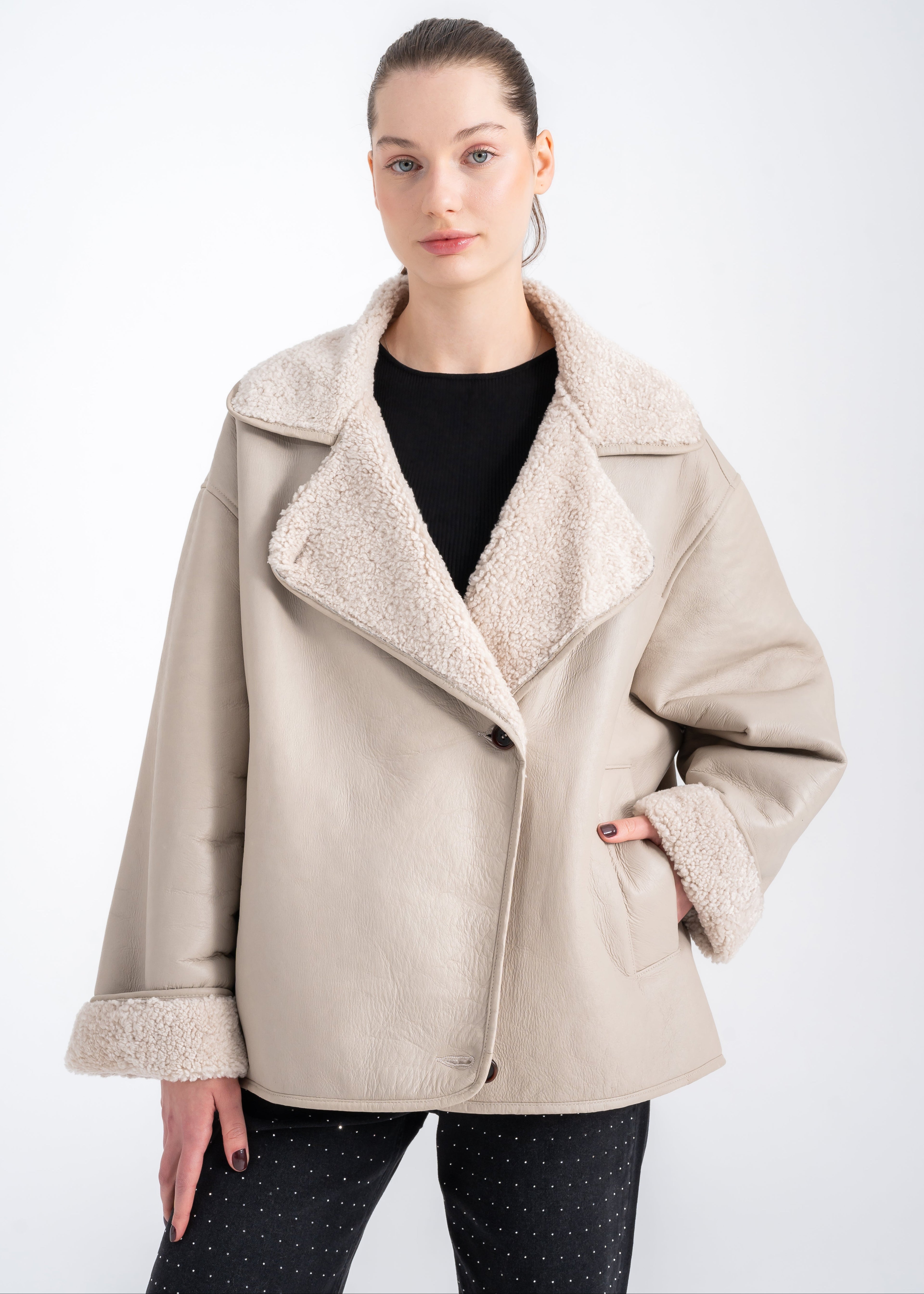 Woman wearing a relaxed light beige shearling jacket with wide lapels, turned up shearling cuffs and front welt pockets by Lebeni.