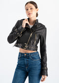 Woman wearing a black lambskin cropped biker jacket with asymmetric gold zipper, eyelet belt, belted cuffs and red lining, styled with high waist blue jeans by Lebeni.