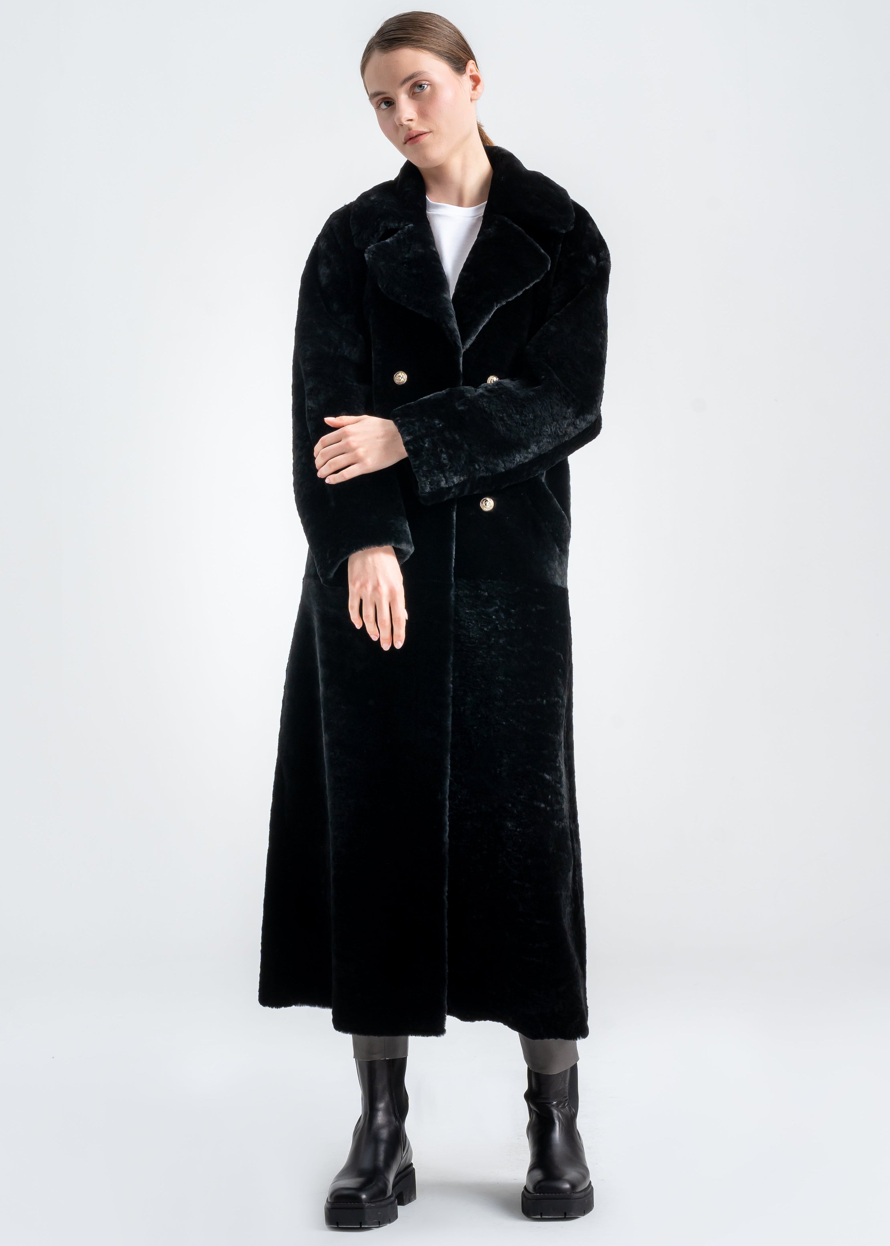 Model wearing a long black shearling coat with wide notch collar, double breasted gold tone buttons, and side pockets by Lebeni.