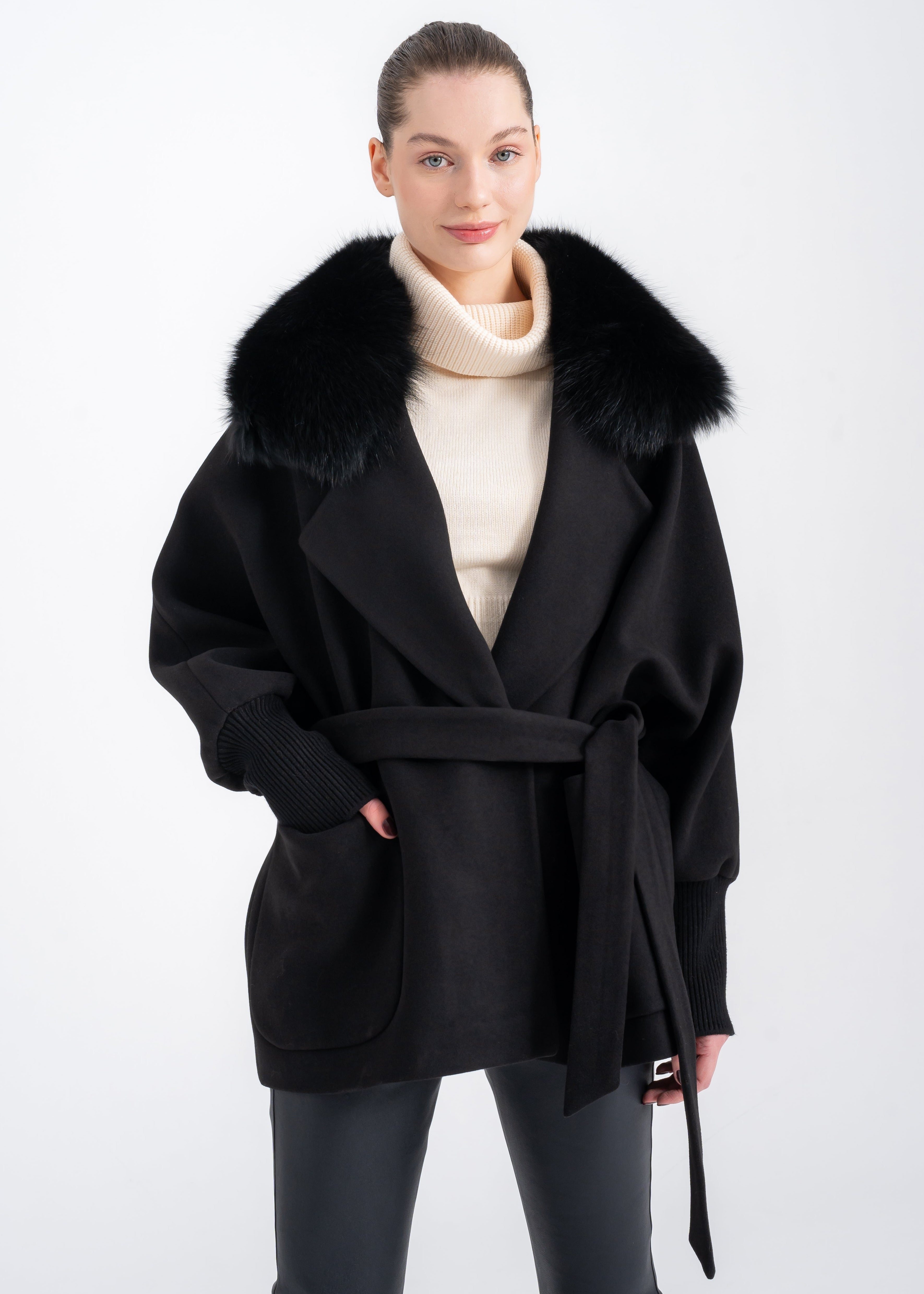 Woman wearing a black wool and cashmere coat with oversized detachable black fur collar, ribbed knit cuffs, wrap belt and front patch pockets by Lebeni.