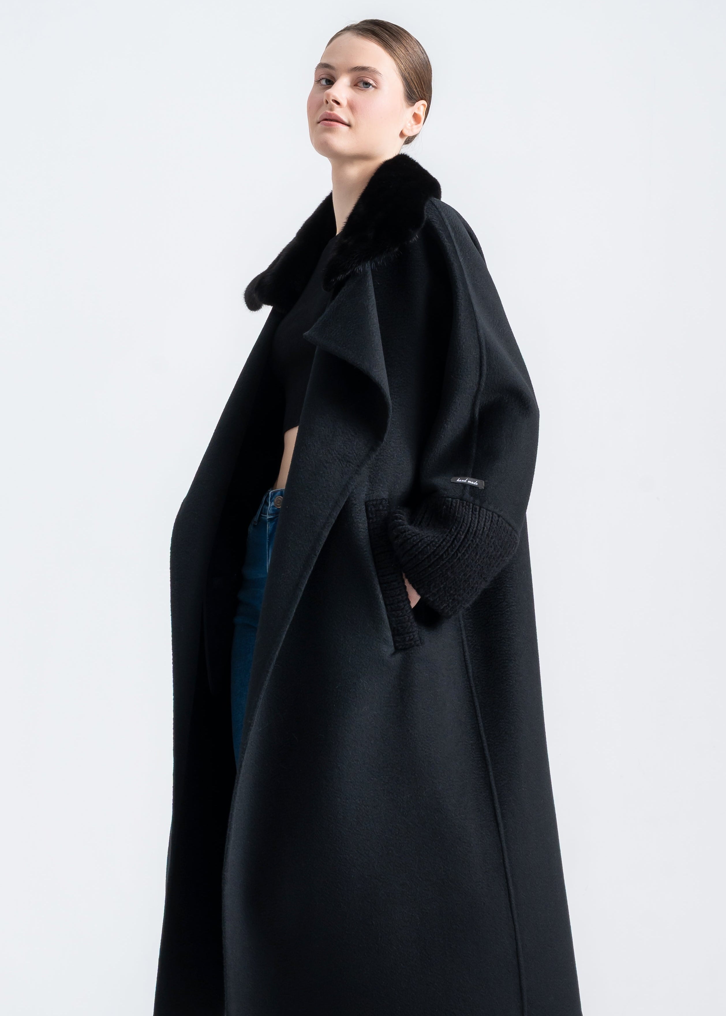 Woman wearing a long black wool cashmere coat with real black mink fur shawl collar, wrap belt, ribbed knit cuffs and knit trimmed front pockets by Lebeni.
