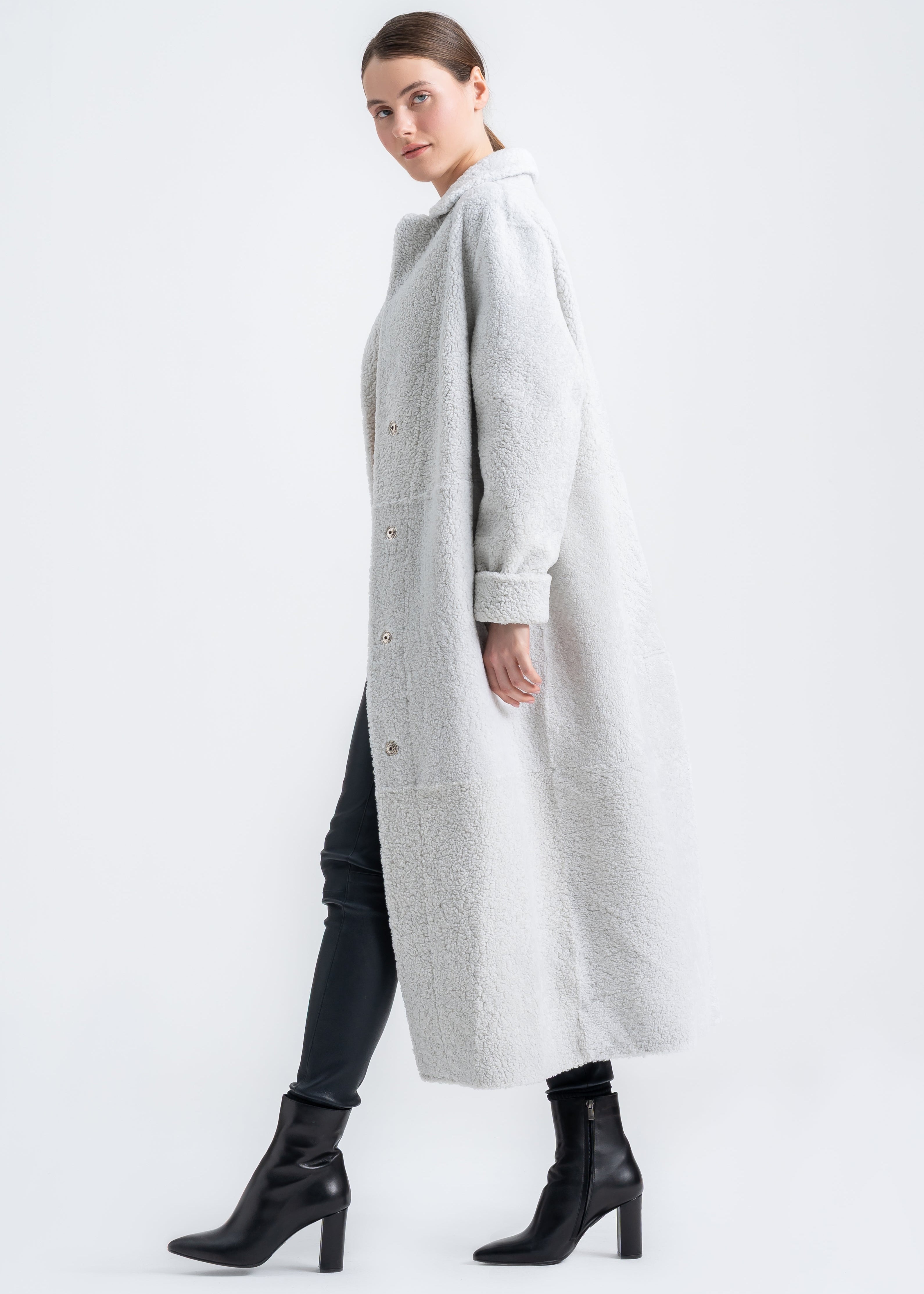 Woman wearing a light grey long shearling coat with notch collar, front button closure and relaxed full length silhouette by Lebeni.