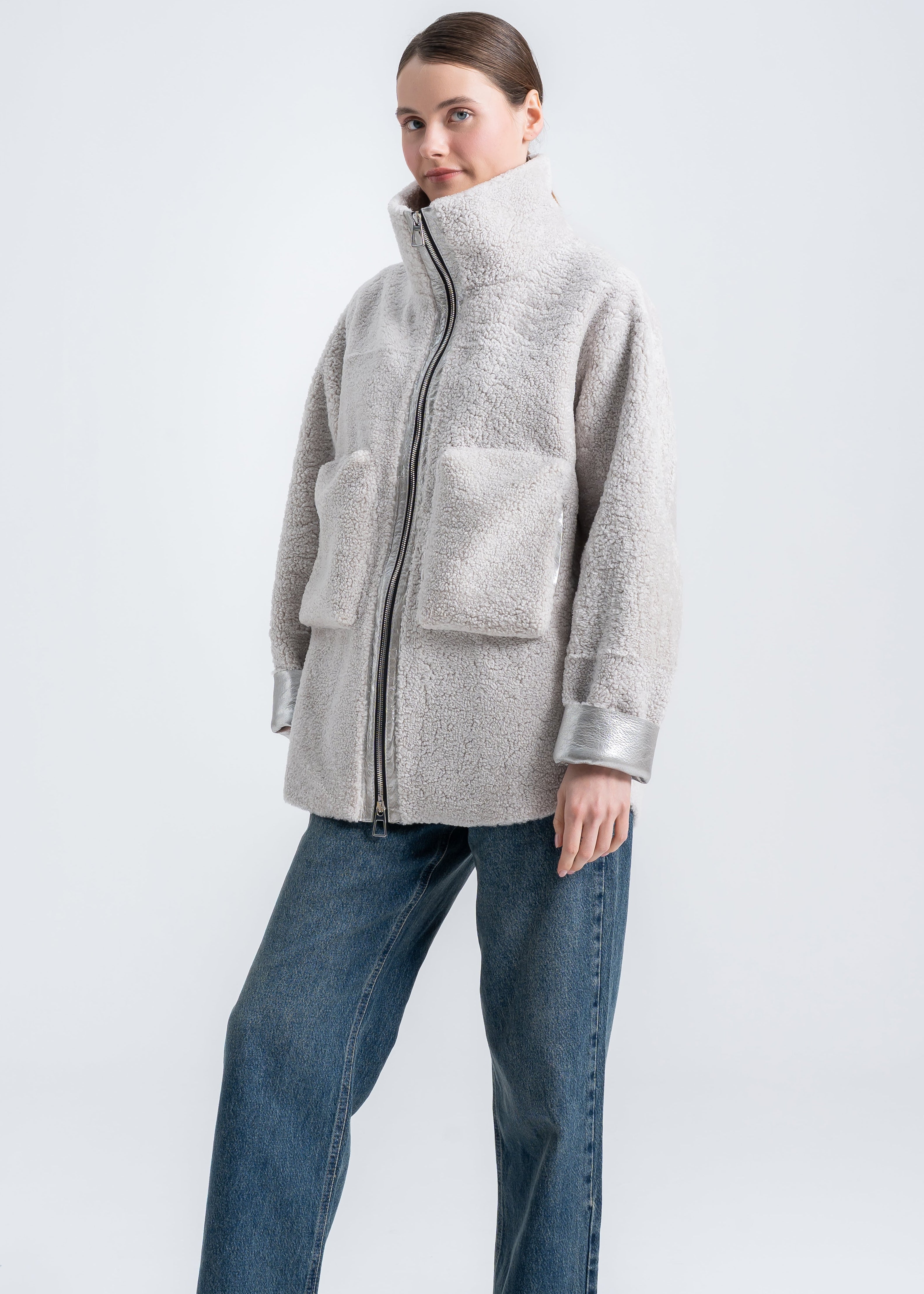Woman wearing a light grey shearling jacket with high funnel collar, front zipper, oversized patch pockets and metallic cuff detail by Lebeni.