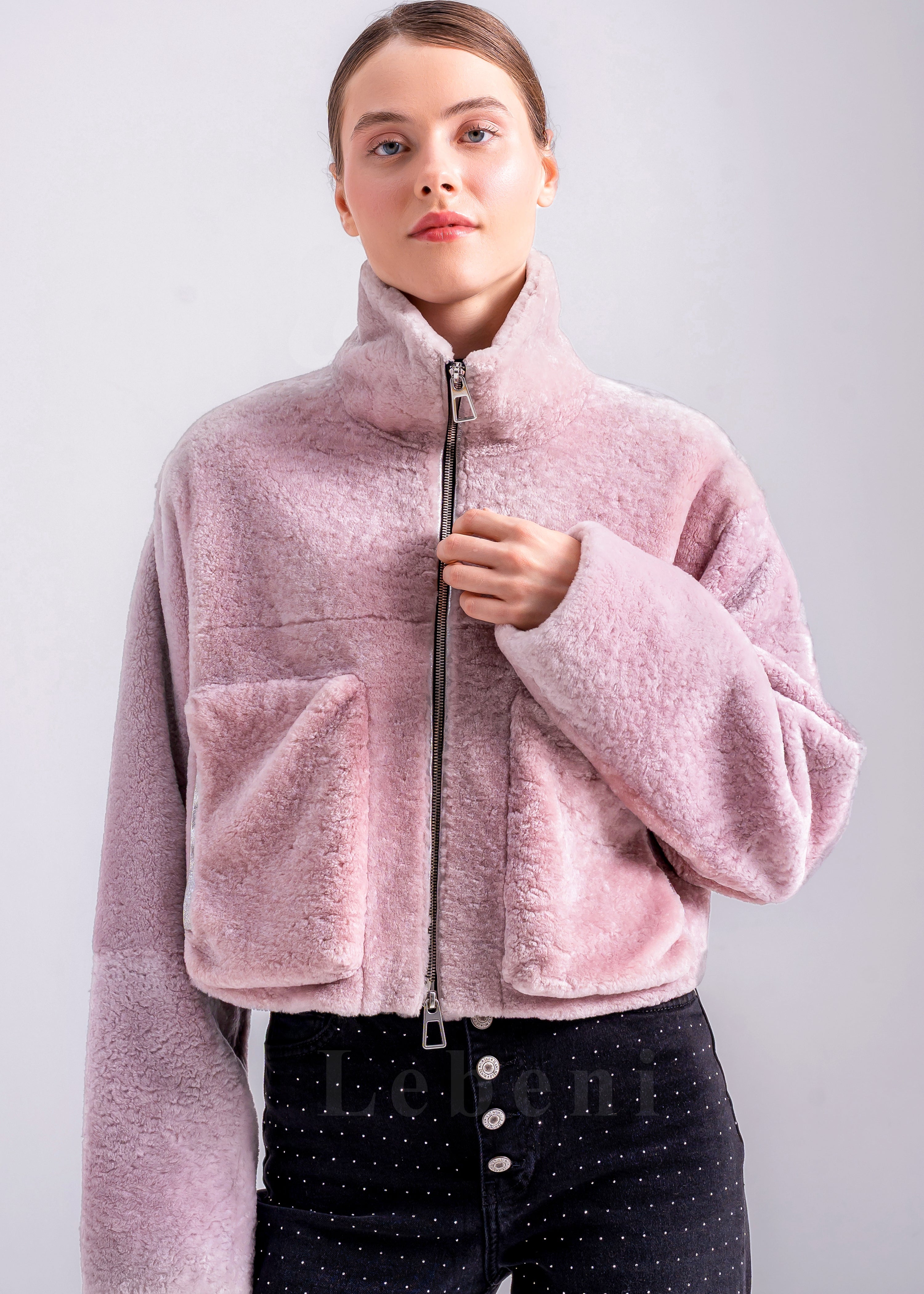 Woman wearing a light pink cropped shearling jacket with high stand collar, front zipper and oversized patch pockets by Lebeni.