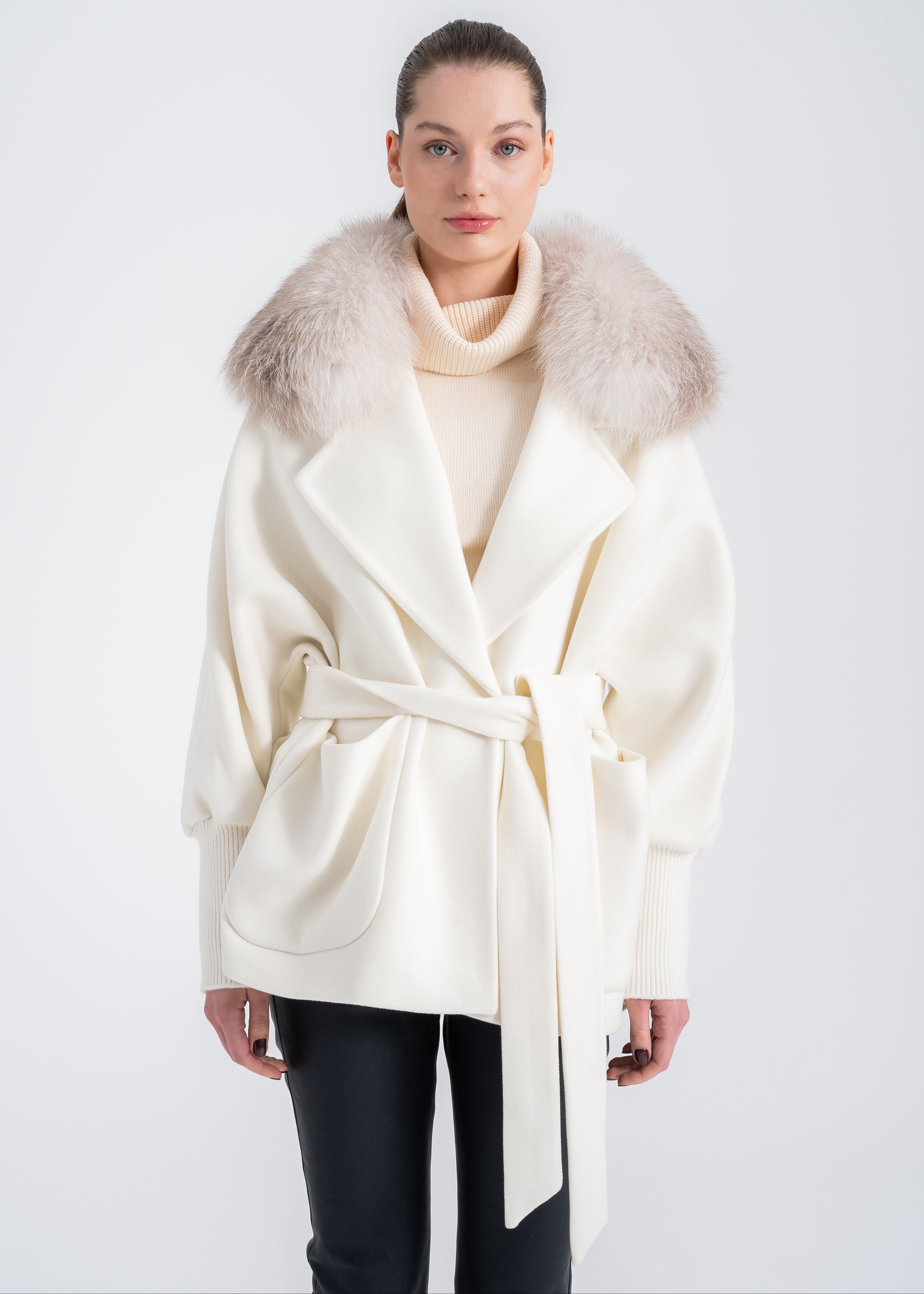 Woman wearing an off white wool and cashmere coat with detachable light beige fur collar, wrap belt, ribbed knit cuffs and curved front patch pockets by Lebeni.