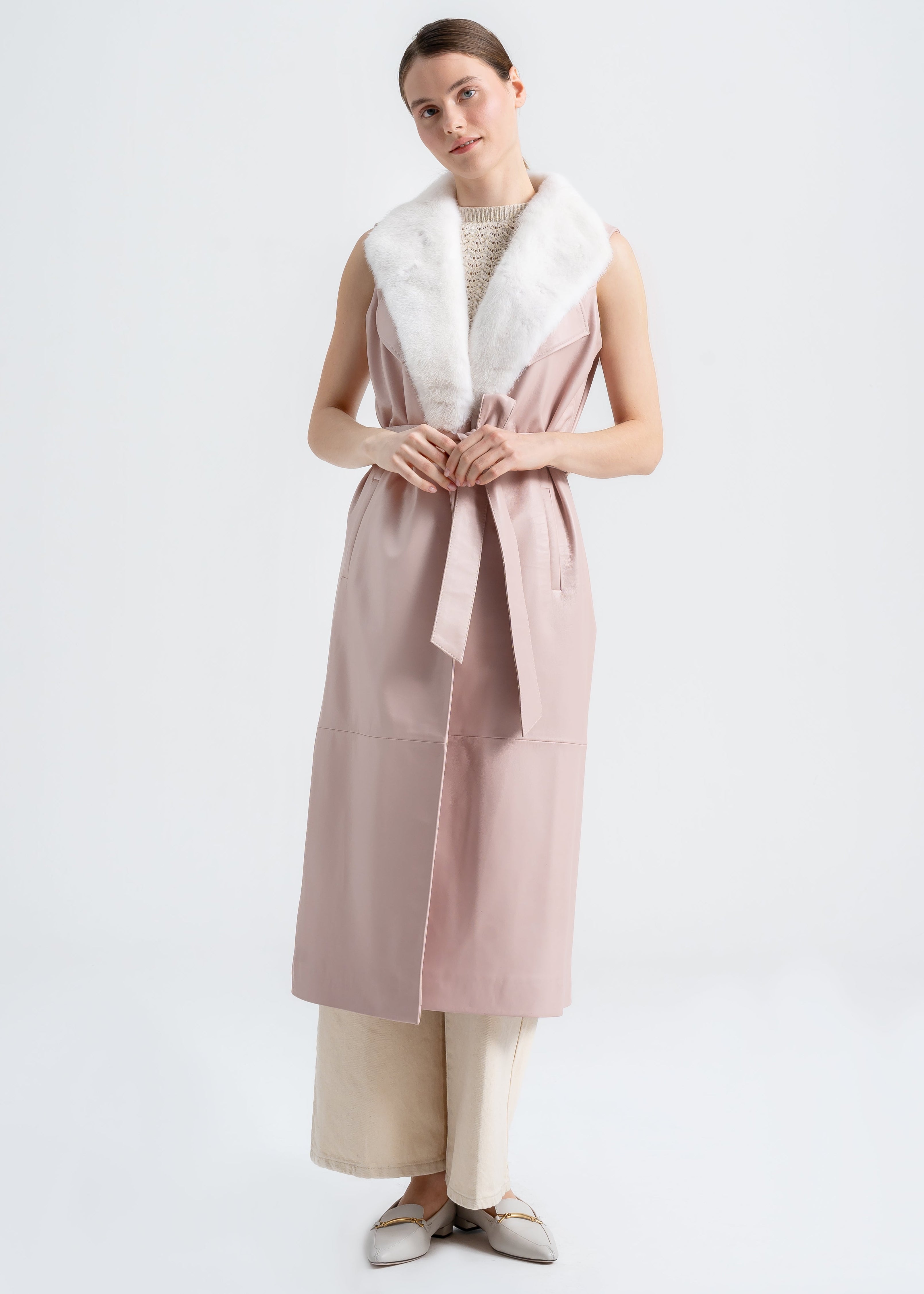 Model wearing a blush lambskin long vest with white mink shawl collar, belted waist, and side pockets by Lebeni.
